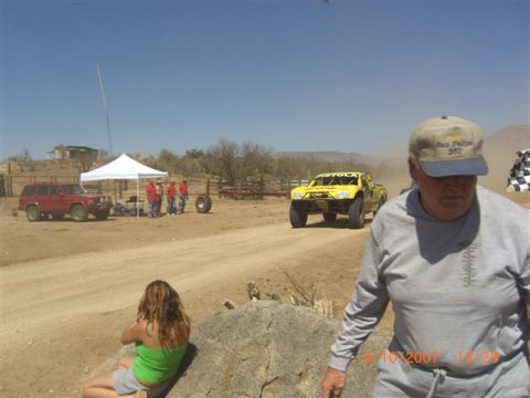 1990 Ford Bronco - Yellow tube-frame off-road race truck speeds along a dusty desert course past a man in a cap and seated spectators.