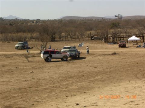 1990 Ford Bronco - Trucks and SUVs gather at a dusty desert staging area with a checkered flag, white tents and a windmill on the horizon.