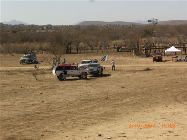 Vehicles gathered at a dusty desert staging area with windmill and tents