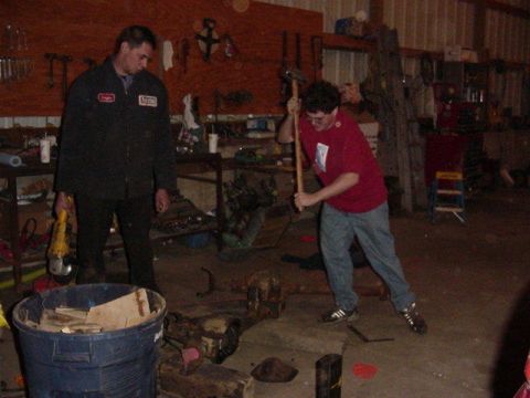 1992 Ford Bronco - A workshop scene with two men engaged in activity, one holding a tool and the other preparing to strike an object with a sledgehammer.