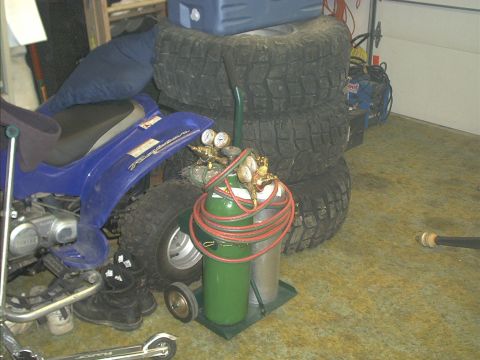 1986 Ford Bronco - A blue ATV sits beside a stack of large tires and a welding tank in a garage.