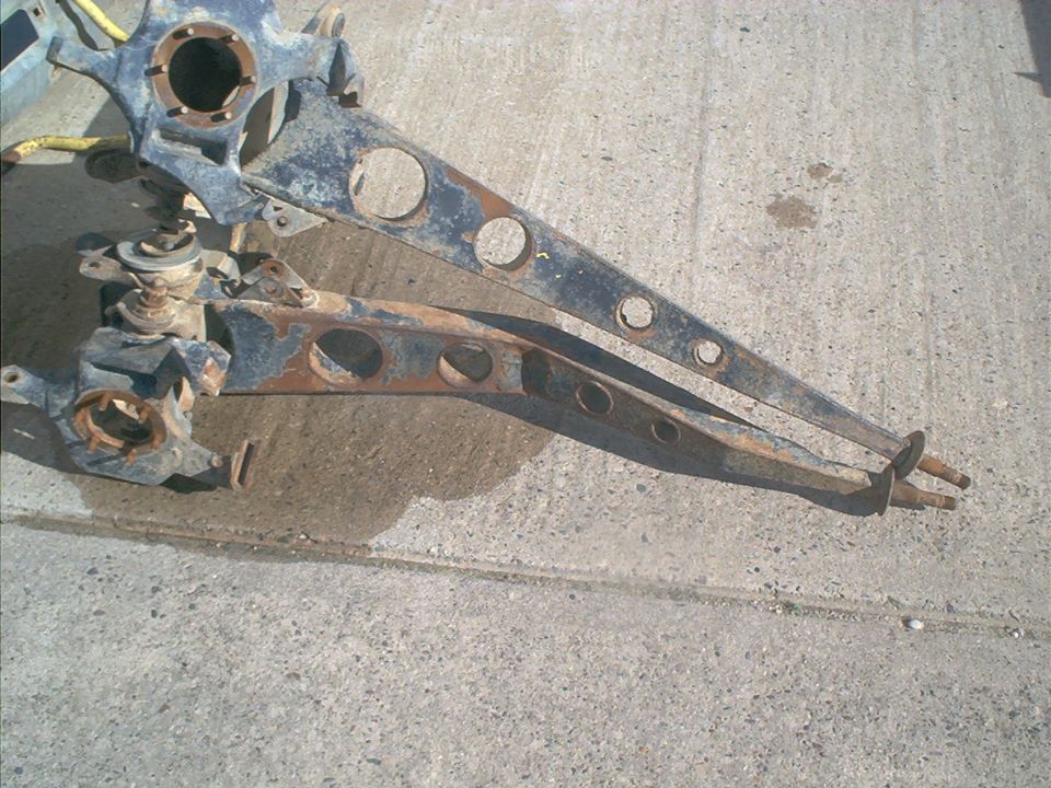 Pair of rusty, dark-colored front axle suspension arms with circular cutouts, resting on a concrete surface