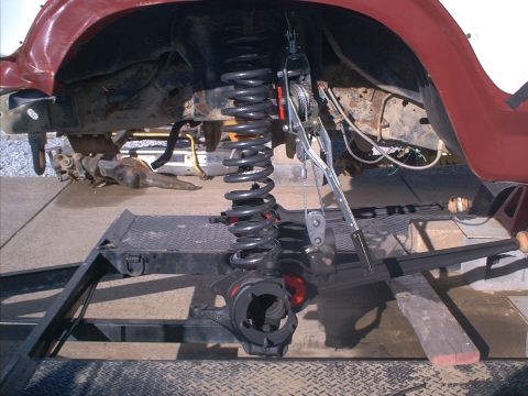 1986 Ford Bronco - Front suspension and axle assembly on a trailer, with a coil spring being compressed by a specialized tool.