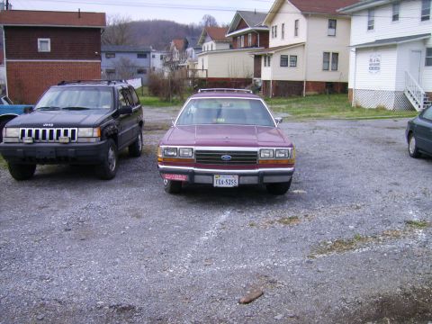 1982 Ford LTD Crown Victoria - Maroon Ford full-size sedan photographed head-on in a gravel lot beside a black Jeep Grand Cherokee, with clapboard houses and a wooded hil…