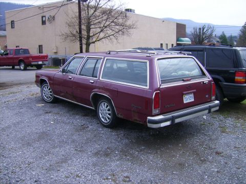 1982 Ford LTD Crown Victoria - Maroon Ford LTD Crown Victoria station wagon shot from the driver-side rear 3/4 in a gravel lot, with rust perforation along the lower quar…