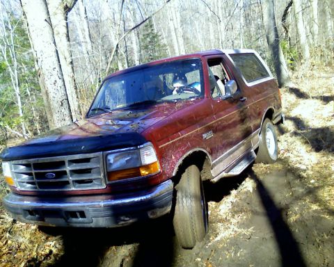 1996 Ford Bronco - A red Ford Bronco navigates a muddy, wooded path, with its tires deep in the dirt.