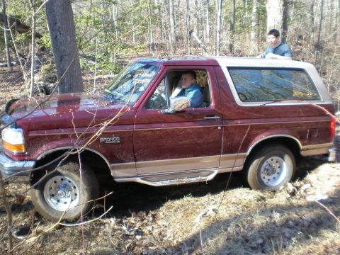 1996 Ford Bronco - A maroon and tan Ford Bronco navigates rough terrain with two people inside and on top.