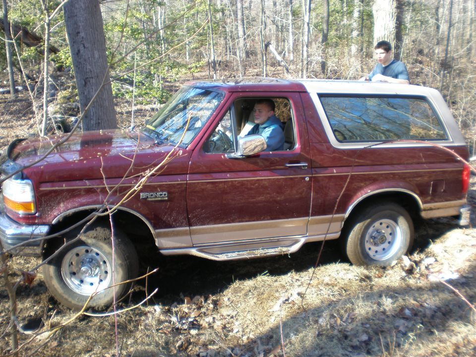 Maroon and tan Ford Bronco off-road, driver visible, with a person standing on the roof
