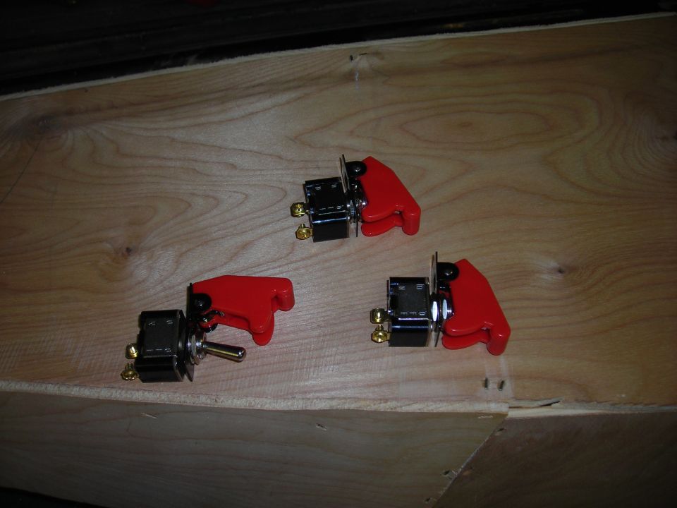 Three toggle switches with red protective covers are laid out on a wooden surface