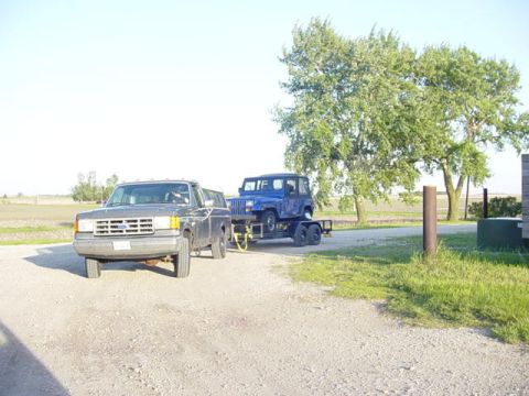 1992 Ford Bronco - A gray Ford Bronco is parked on a gravel surface, towing a blue Jeep Wrangler on a trailer.