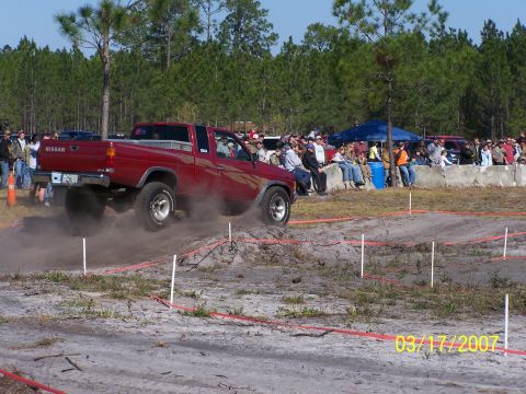 1989 Ford Bronco - A red Nissan pickup truck navigates an off-road course, with spectators watching from the sidelines.