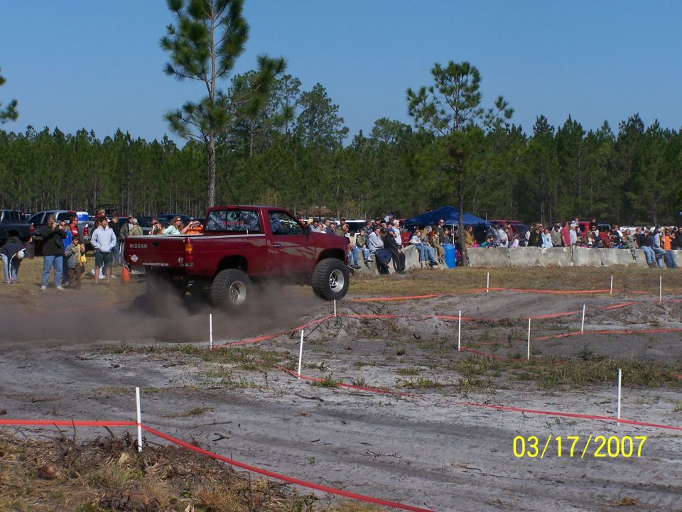 Red pickup truck with lifted suspension kicking up dust on an off-road track, crowd watching in background