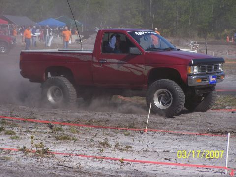 1989 Ford Bronco - A lifted red pickup truck navigates a dusty off-road course with onlookers present.