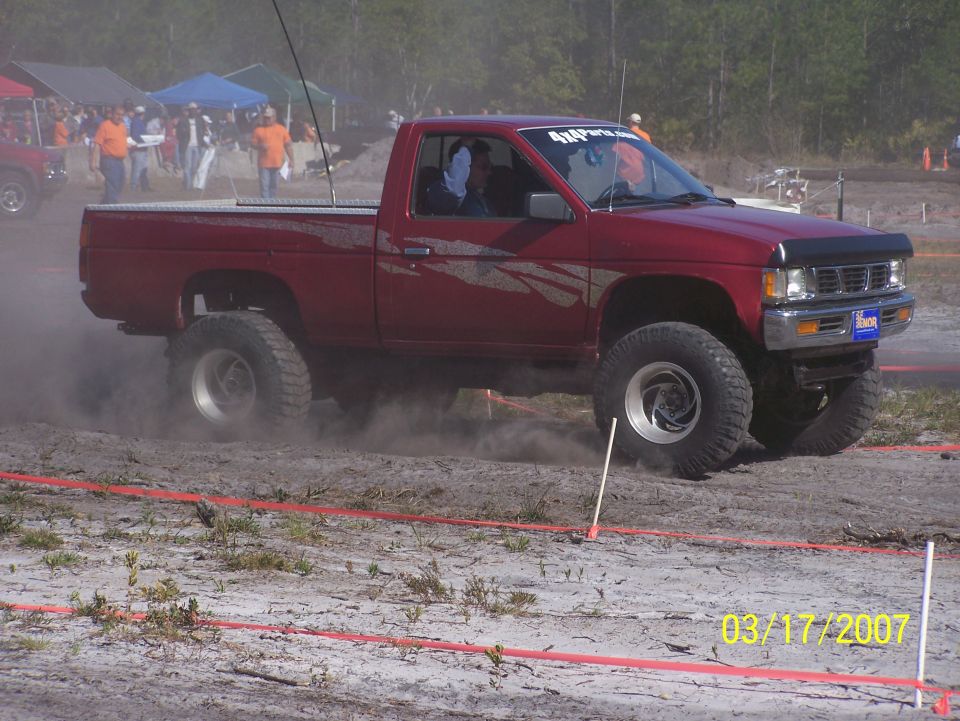 Red pickup truck with lifted suspension kicking up dust on a dirt track. Another red vehicle is visible in the background with spectators
