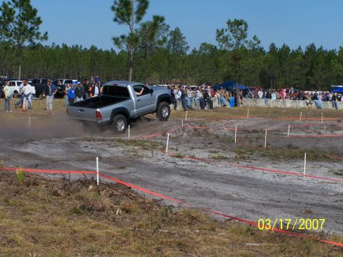 1989 Ford Bronco - A silver pickup truck navigates a dirt track during an event, with spectators watching from the sidelines.