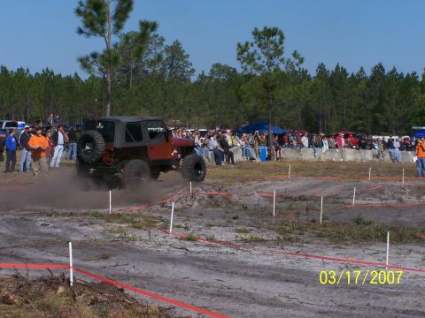 1989 Ford Bronco - A red Jeep Wrangler navigates an off-road course, kicking up dust as spectators watch from the sidelines.