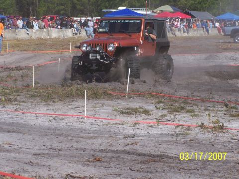 1989 Ford Bronco - Jeep Wrangler navigates a muddy off-road course with a crowd watching.