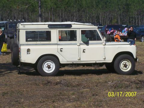 1989 Ford Bronco - A cream-colored Land Rover Series III station wagon is parked outdoors with other vehicles in the background.