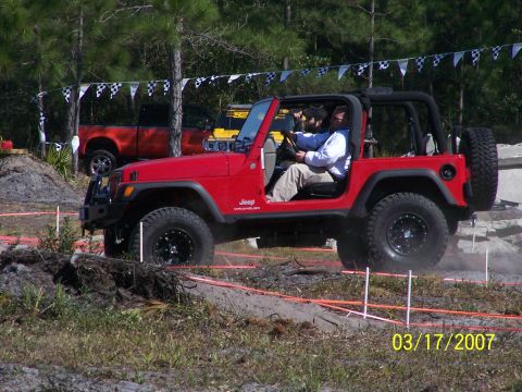 1989 Ford Bronco - Red Jeep Wrangler navigates an off-road obstacle course, with a pickup truck in the background.