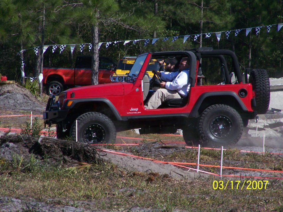 Red Jeep Wrangler driving on an off-road course with two people inside. A red pickup truck is visible in the background