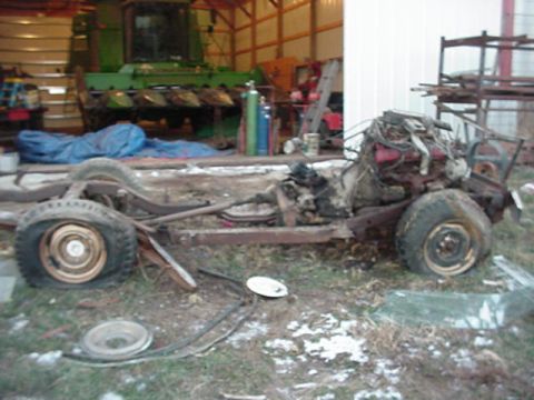 1992 Ford Bronco - A stripped donor truck reduced to a bare rusty frame with engine still mounted, sitting on patchy snow-dusted grass beside a pole barn hous…