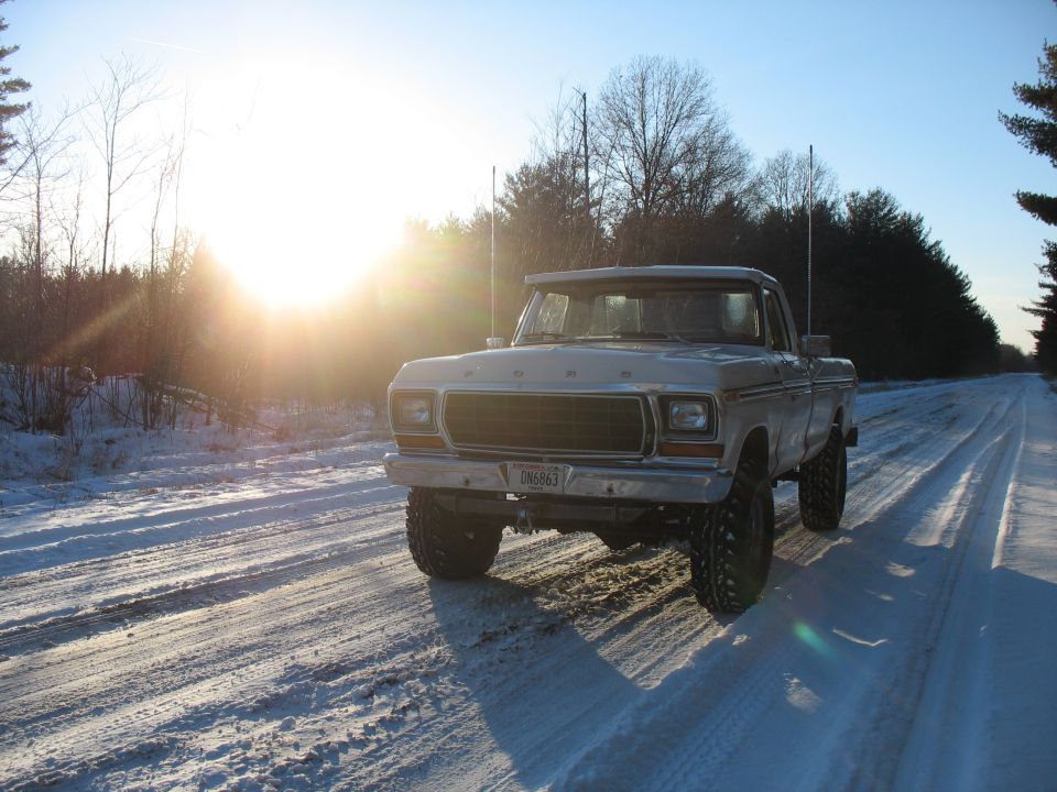 Lifted white Ford pickup on a snowy tree-lined road at sunset, front three-quarter view