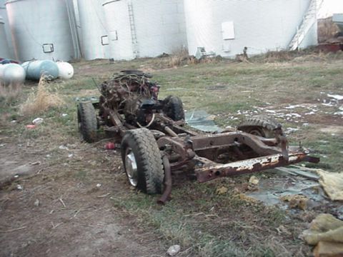 1992 Ford Bronco - A stripped donor truck rolling chassis sits in a weedy lot beside grain bins, with the body removed to expose the rusty ladder frame, leaf…
