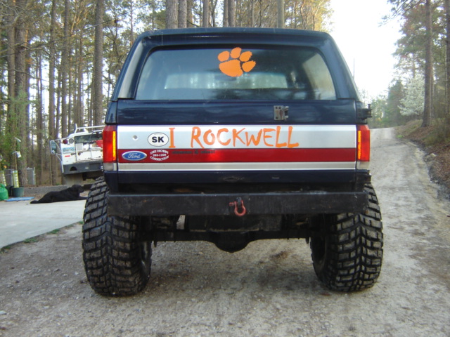 Rear view of a dark blue Ford Bronco with a silver stripe, red stripe, and orange text reading 'I ROCKWELL'. Has a paw print decal on the rear window and a Ford badge