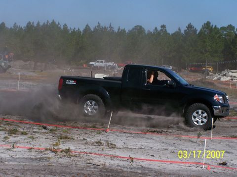 1989 Ford Bronco - A black Nissan pickup truck drifts on a dirt track, creating a cloud of dust. Other vehicles and trees are visible in the background.