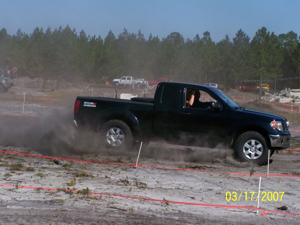 Black Nissan pickup truck kicking up dust while drifting on a dirt track. Another white truck is visible in the background