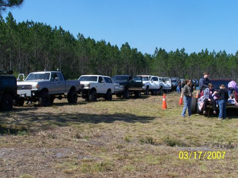1989 Ford Bronco - Several lifted trucks and SUVs are parked in a grassy field with trees in the background.
