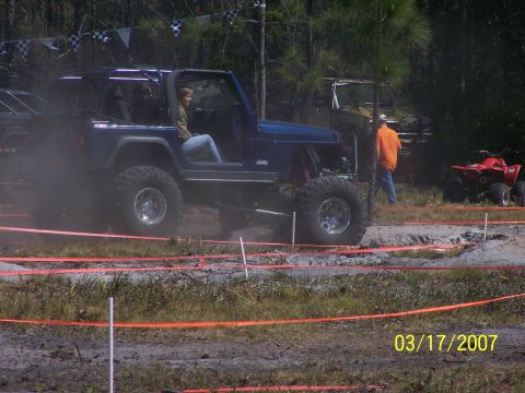 1989 Ford Bronco - A blue Jeep navigates a muddy course with spectators and other vehicles present.