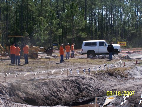 1989 Ford Bronco - A white SUV is positioned on a dirt obstacle course, with a backhoe and several people in the background.