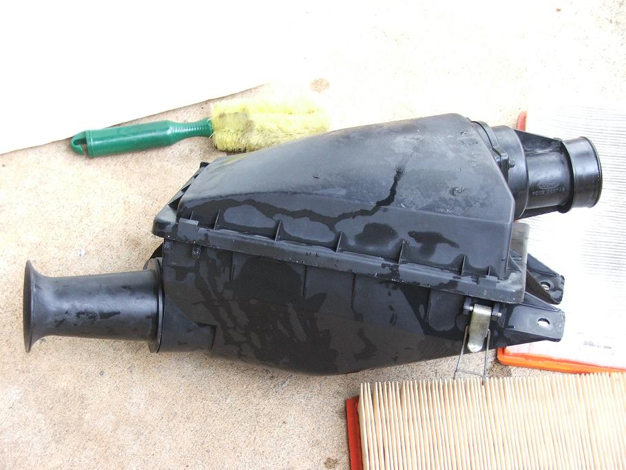 Black plastic air cleaner housing with a green brush and yellow sponge nearby. A new air filter is visible