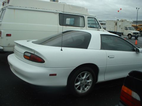 2000 Ford Crown Victoria - A white coupe is parked in a lot next to a white van under a cloudy sky.