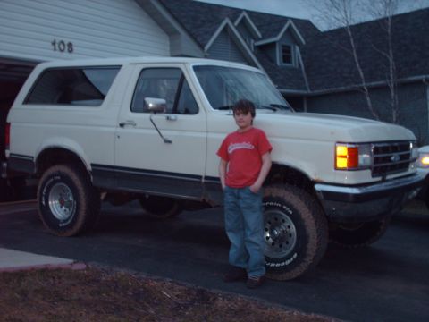 1990 Ford Bronco - A boy stands next to a white 1990 Ford Bronco parked in a driveway.