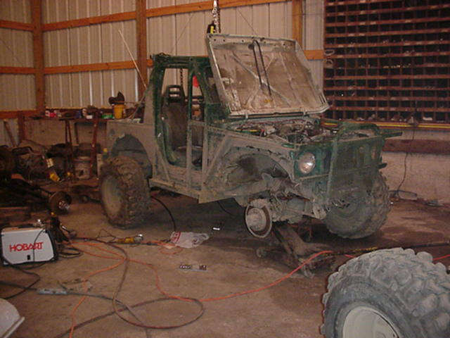 Green off-road vehicle with hood open, missing front fenders and one wheel, in a garage
