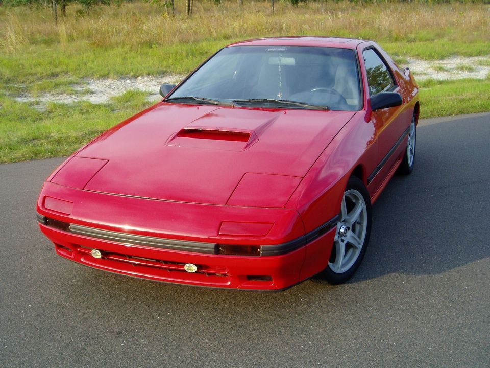 Red Mazda RX-7 with pop-up headlights and a hood scoop, parked on asphalt with grass in the background