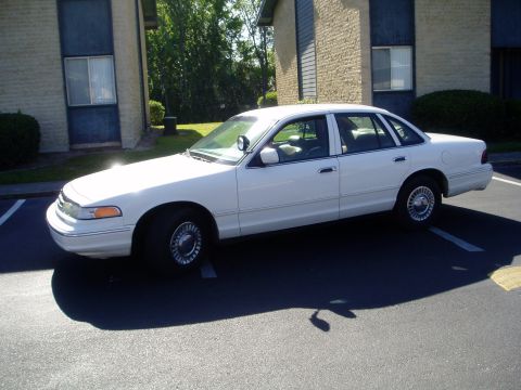 2000 Ford Crown Victoria - A white sedan, likely a 1998 Ford Crown Victoria, is parked in a lot with apartment buildings in the background.