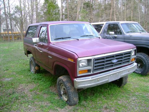 1989 Ford Bronco - Maroon 1989 Ford Bronco sits in a grassy area with flat tires; a dark SUV is parked behind it.