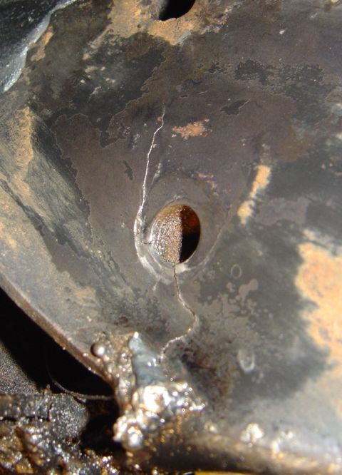 1986 Ford Bronco - Extreme close-up of a rusty black-painted steel bracket or panel with a visible crack running through a bolt hole, the crack traced by a sc…