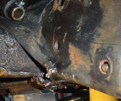 1986 Ford Bronco - Underbody close-up of a grimy steel frame bracket with a visible crack running down through a hole toward the lower edge, where rough weld…
