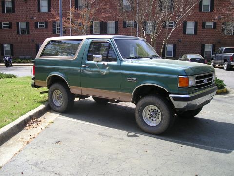 1989 Ford Bronco - A green Ford Bronco with tan trim is parked in a lot with a brick building in the background.
