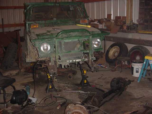Green off-road vehicle on jack stands in a garage, front end disassembled. Spare tires and tools are visible