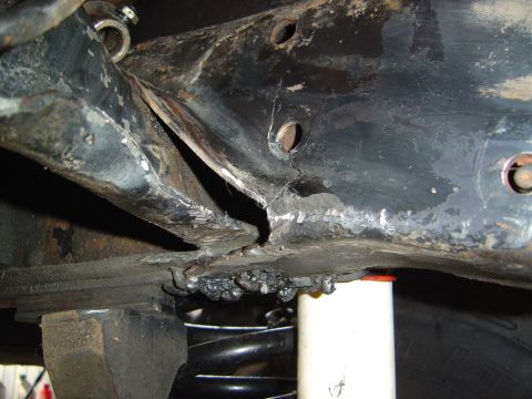1986 Ford Bronco - Underside view of a vehicle frame showing a significant crack and separation in the black-painted steel frame rail, with crude weld beads v…