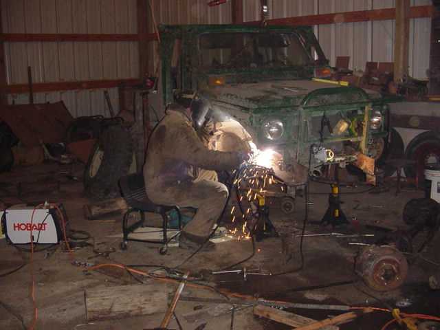 Man in welding gear working on the front end of a green vehicle in a garage. A Hobart welder is visible
