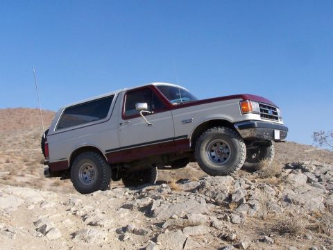 1990 Ford Bronco - Side profile of a two-tone burgundy-and-white full-size Ford Bronco with removable hardtop parked on a rocky desert hillside, front wheels…