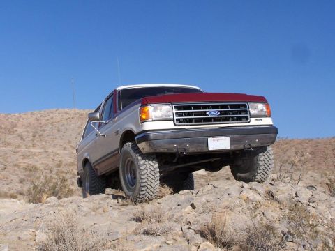 1990 Ford Bronco - Low-angle three-quarter front view of a two-tone red-over-tan Ford Bronco climbing a rocky desert hillside against a clear blue sky, front…