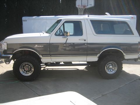 1989 Ford Bronco - Driver-side profile of a lifted white-over-dark two-tone Ford Bronco with hardtop, parked on a concrete driveway with a white enclosed trai…