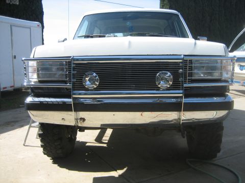 1989 Ford Bronco - Head-on view of a white lifted full-size Ford truck on a sunny concrete pad, fitted with a chrome tube grille guard/brush bar carrying two…